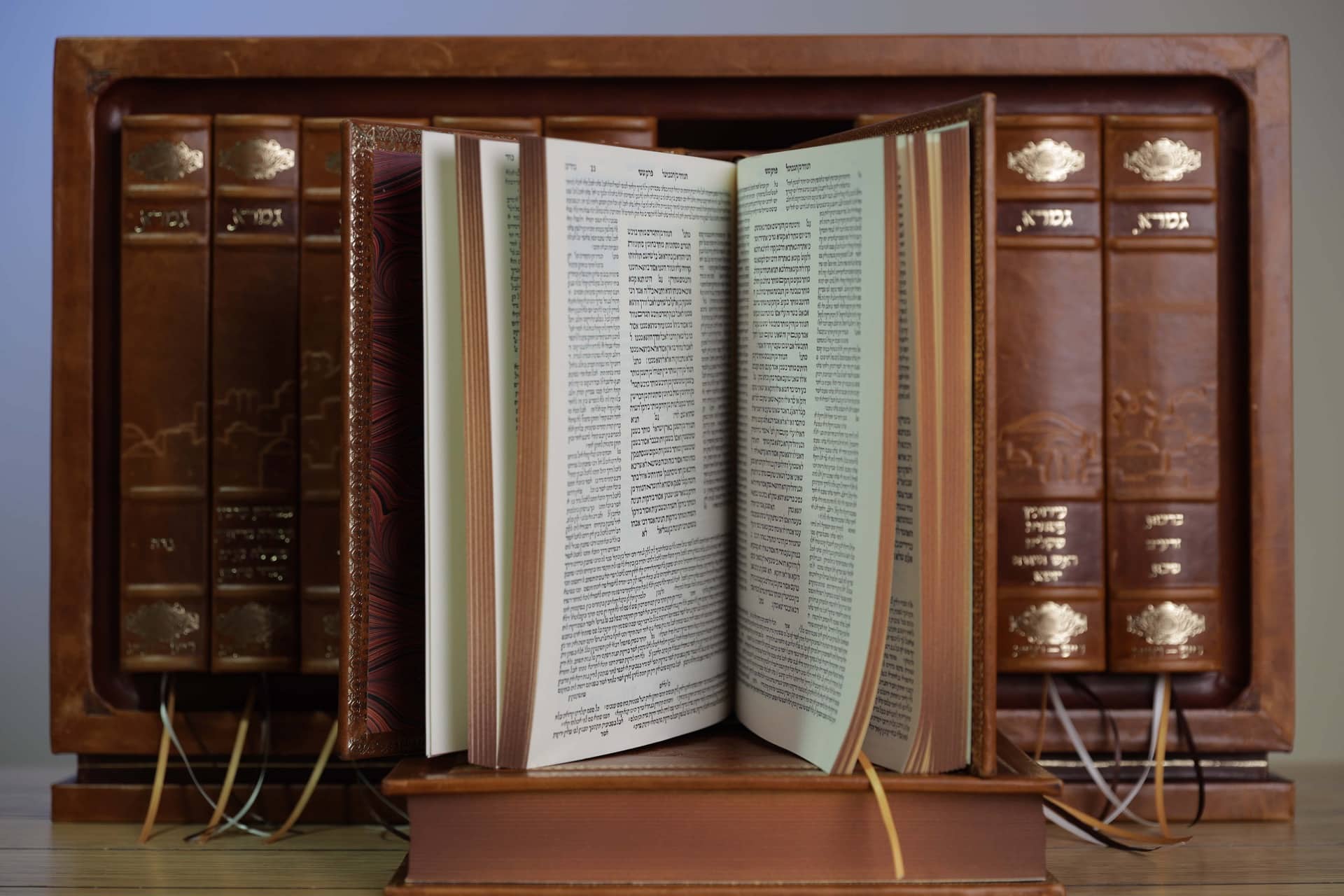 An open gemara on a stand, surrounded by leather-bound seforim with gold-stamped spines